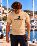 Modelo masculino con camiseta SPORT STYLE de IMPARABL3 en color arena 07, modelo Roly Stafford de corte regular, 100 % algodón de 190 g/m². Logotipo I3 IMPARABL3 negro centrado en el pecho, fotografiado junto a un puerto deportivo.