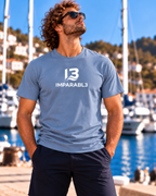 Modelo masculino con camiseta SPORT STYLE Azul Zen de IMPARABL3, confeccionada en algodón de 190 g/m² y corte regular. Logotipo I3 IMPARABL3 blanco centrado en el pecho. Fotografía junto a un puerto deportivo con veleros y el mar de fondo.