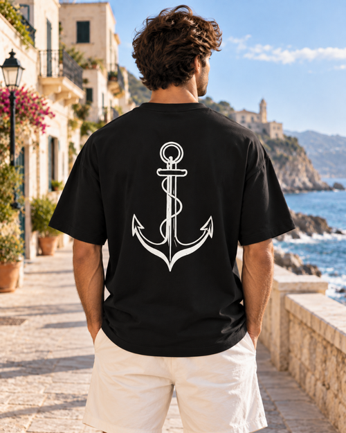 Hombre con camiseta oversize negra de IMPARABL3, vista trasera con gran diseño de ancla blanco junto al mar.