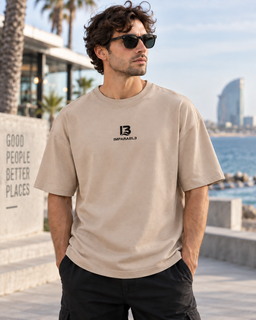 Modelo masculino con camiseta oversize de IMPARABL3 en color arena 07, mostrando el logo frontal en un entorno urbano junto al mar