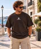 Modelo masculino con camiseta oversize de IMPARABL3 en color ébano 231, mostrando el logo frontal blanco en un entorno urbano junto al mar