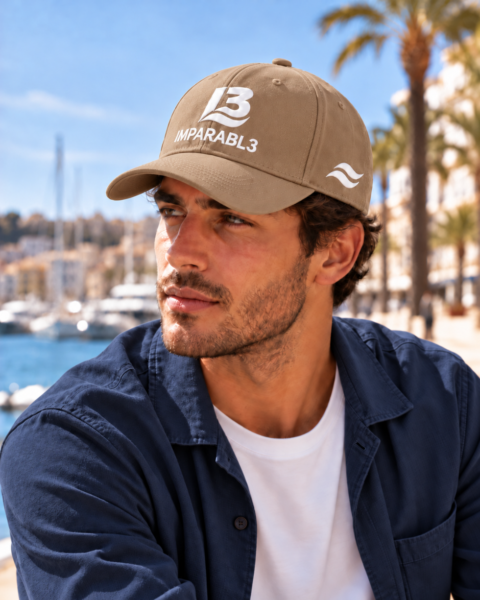 Hombre con gorra beige de IMPARABL3, color 12, logotipo I3 blanco y dos olas blancas en el lateral izquierdo, combinada con sobrecamisa azul marino en un puerto mediterráneo.