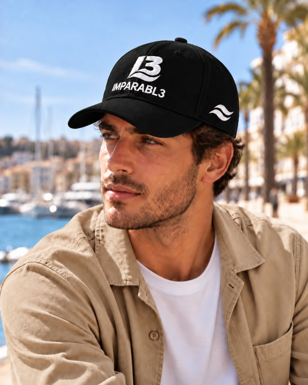 Hombre con gorra negra de IMPARABL3, logotipo I3 blanco y dos olas blancas en el lateral izquierdo, combinada con sobrecamisa beige en un puerto mediterráneo.