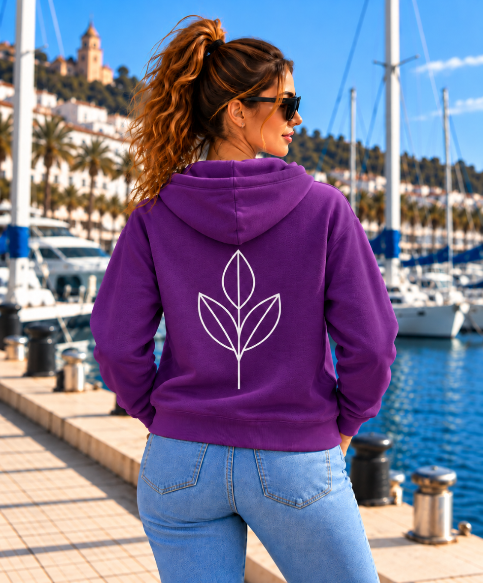 Mujer de espaldas con sudadera SAVIA púrpura IMPARABL3, con capucha, cremallera y diseño botánico blanco en la espalda, junto a un puerto deportivo.