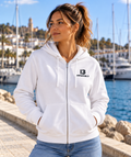 Modelo con sudadera blanca ALBORADA de mujer de IMPARABL3, con capucha, cremallera frontal y logo I3 negro en el pecho, junto a un puerto mediterráneo.