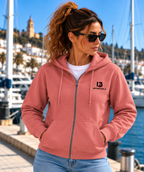 Mujer con gafas de sol luciendo la sudadera IMPULSO de IMPARABL3 en color rosatón, con capucha, cremallera y logo I3 negro, junto a un puerto mediterráneo.