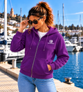 Mujer con sudadera IMPULSO púrpura 71 de IMPARABL3, con capucha, cremallera metálica y logo I3 blanco en el pecho, junto a un puerto deportivo.